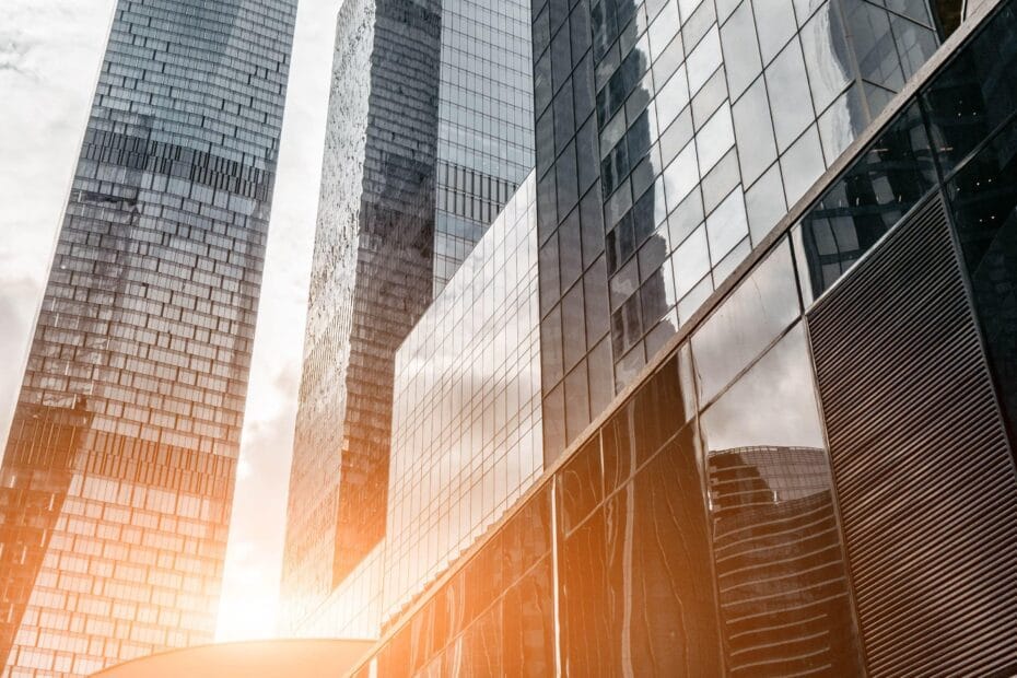 Vue dramatique vers le ciel de gratte-ciels modernes en verre aux façades réfléchissantes capturant la lumière dorée du coucher du soleil. Les bâtiments présentent des détails architecturaux distinctifs, notamment des éléments courbés et des motifs géométriques, sous un ciel nuageux.