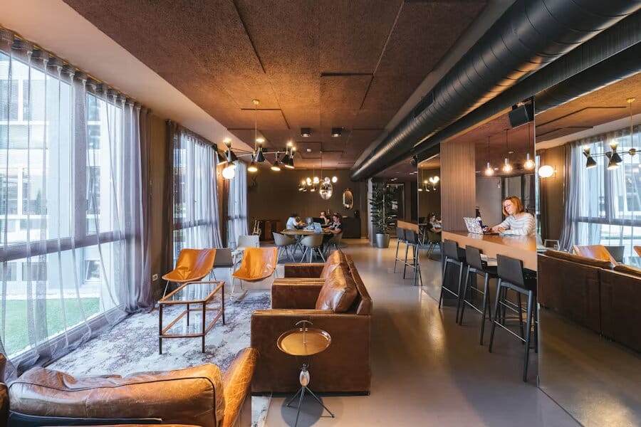 Un espace de coworking spacieux avec des éléments de design industriel, notamment des conduits apparents, de grandes fenêtres avec des rideaux légers, des canapés en cuir, un éclairage suspendu élégant et une zone de travail à hauteur de bar. L’espace allie confort et fonctionnalité avec des tons chaleureux de cuir.