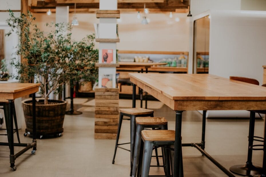 Espace de coworking cosy avec tables en bois, tabourets industriels, plantes et ambiance décontractée.