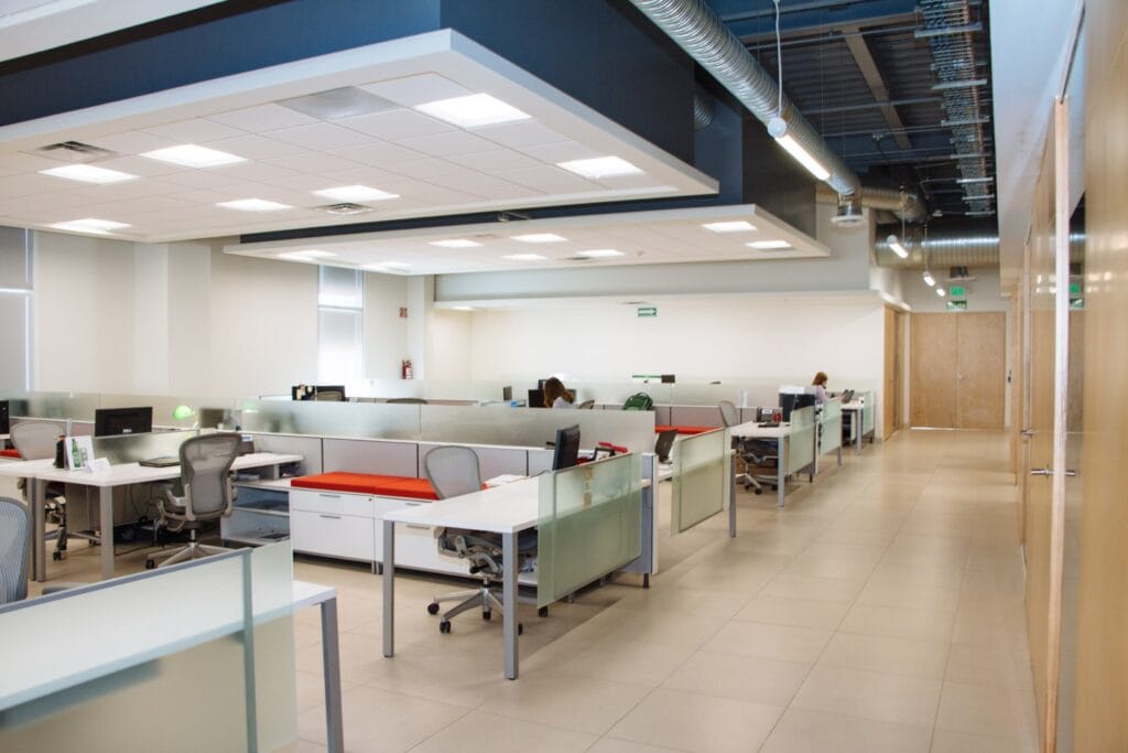Open space lumineux avec plusieurs postes de travail, séparations en verre dépoli, mobilier blanc et orange, et plafond industriel apparent.